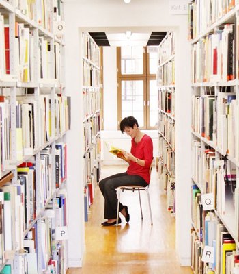 lesende Frau sitzt zwischen zwei deckenhohen Bücherregalen
