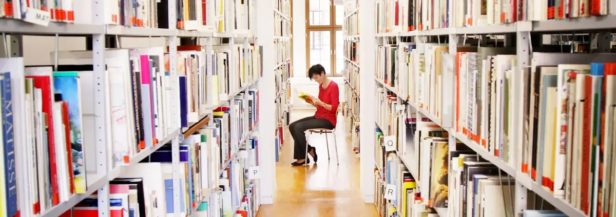 lesende Frau sitzt zwischen zwei deckenhohen Bücherregalen