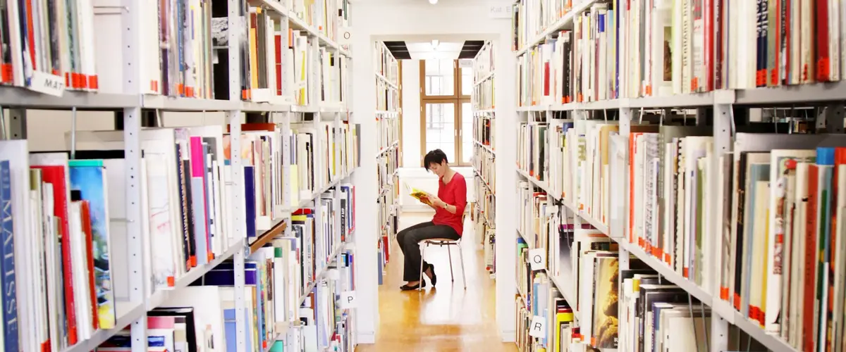 lesende Frau sitzt zwischen zwei deckenhohen Bücherregalen