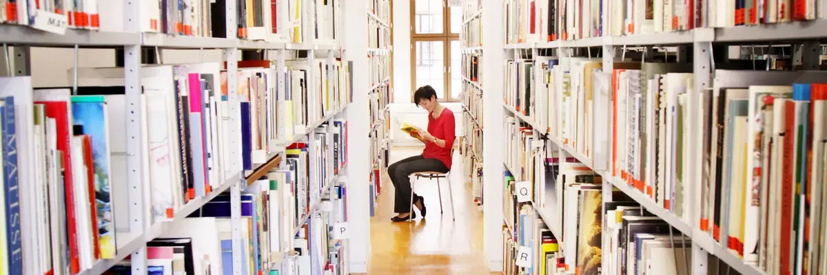 lesende Frau sitzt zwischen zwei deckenhohen Bücherregalen